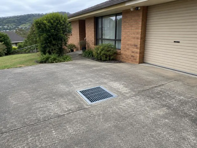 Concrete driveway with a metal drain grate beside a brick house and garage in Bolwarra Heights
