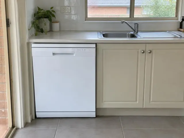 White dishwasher under a kitchen bench beside a stainless sink in a tiled home kitchen in Woodville